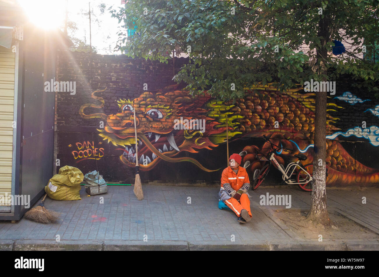Einen chinesischen Arbeiter sitzt vor einer Wand Malerei zeigt einen Drachen auf einer Straße im Stadtteil Chenghua, Chengdu City, im Südwesten Chinas Sichuan pro Stockfoto