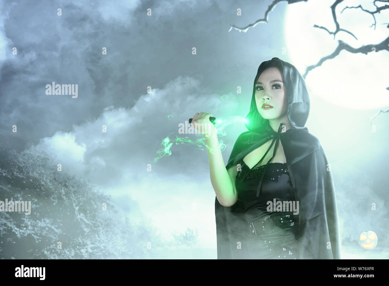 Asiatische Hexe Frau in Schwarz mit Kapuze tun Ritual Magic mit einem Messer mit moonlight Hintergrund. Halloween Konzept Stockfoto