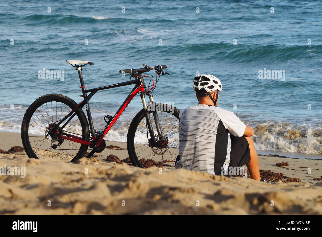 Ein Mann mit einem Fahrrad ist Ruhe am Strand des Atlantik. Sitzt im Sand. Ansicht von hinten. Stockfoto