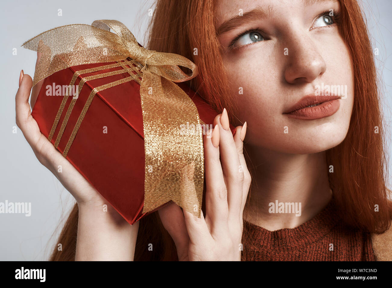 Studio shot von niedlichen und junge rothaarige Frau mit einem Geschenk in ihr Ohr, während sich gegen grauen Hintergrund. Geburtstag Konzept. Feiertage Stockfoto