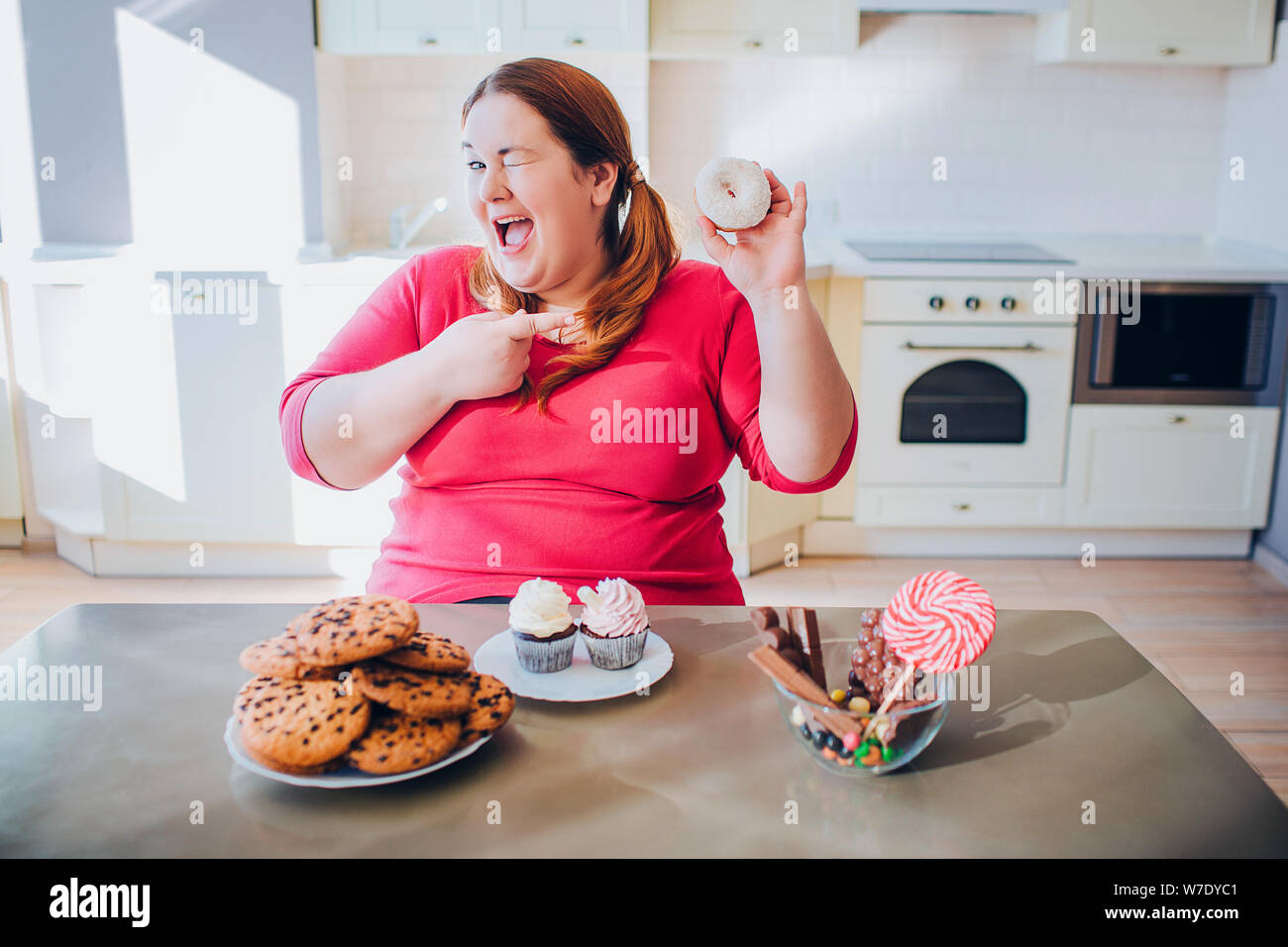 Fett junge Frau in der Küche sitzt und isst süße Speisen. Happy plus größe Modell Lächeln auf Kamera und Punkt auf Donut. Tageslicht in der Küche. Cookies und p Stockfoto