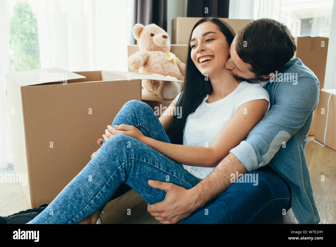 Reizender Mann küsste seine Frau, während auf dem Boden im neuen Zuhause sitzen. Glückliches Paar Umzug, neues Leben in die neue Wohnung Stockfoto