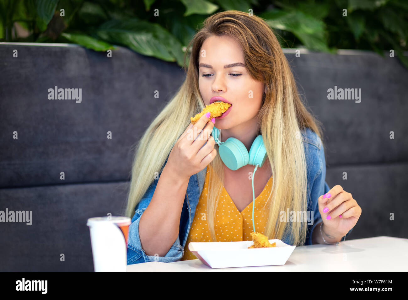 Junge lächelnde trendy Frau essen knusprige Streifen an Fast Food - Happy Hippie girl legere Kleidung und Headsets holding Fried Chicken Stockfoto