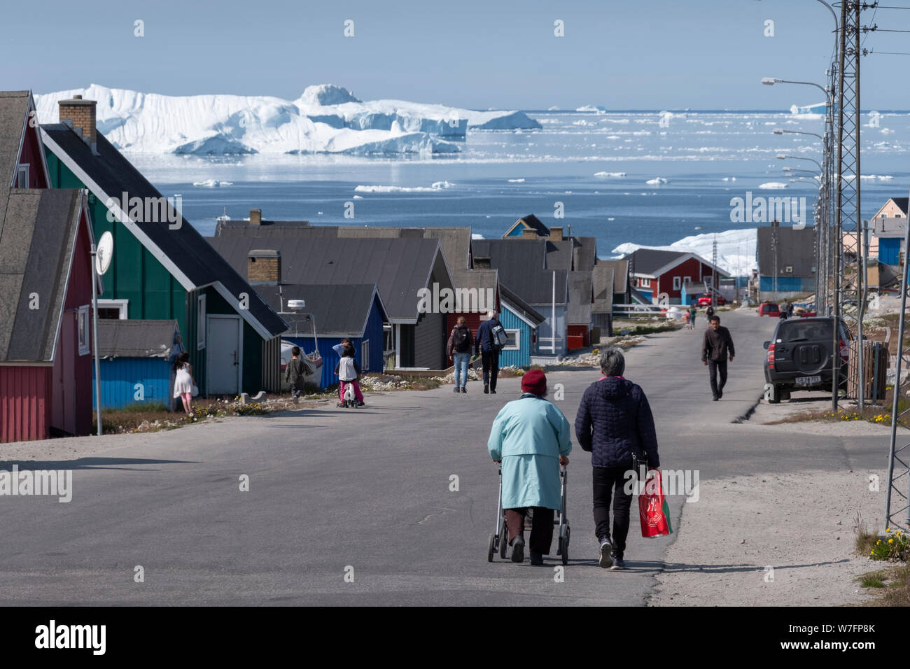 Ältere Menschen zu Fuß auf einer Straße in Ilulissat, Grönland, mit riesigen Eisberge im Hintergrund. Stockfoto