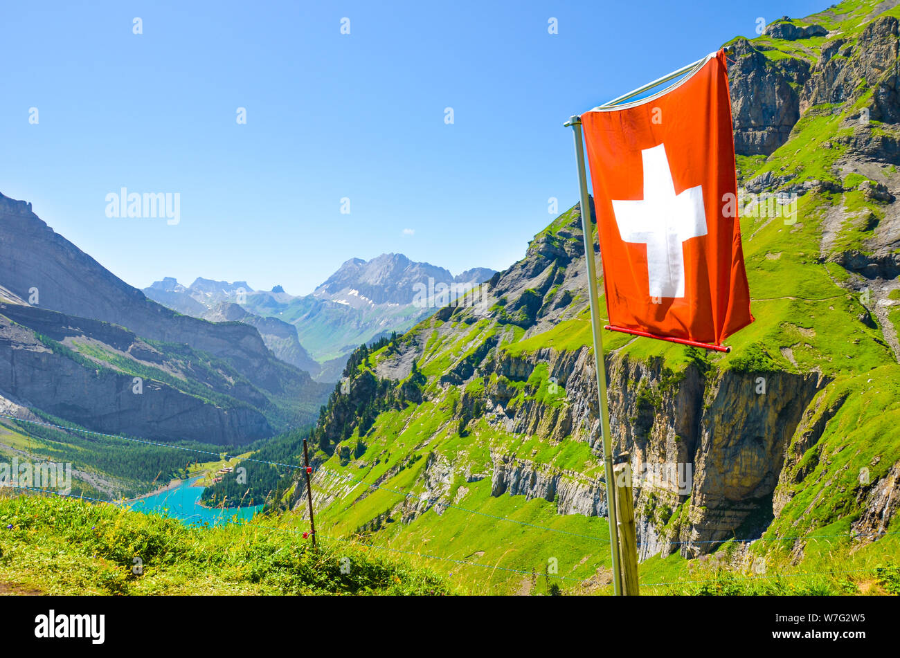 Wehende Flagge der Schweiz auf dem Hügel in den Schweizer Alpen. Türkis Oeschinensee im Hintergrund. Nationales Konzept. Schweiz Sommer. Alpine Landschaft. Meisterwerke der Natur. Touristische Attraktionen. Stockfoto