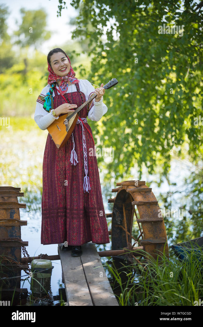 Junge lächelnde Frau in traditionellen russischen Kleidung steht auf einer kleinen Brücke in der Nähe des Sees, und spielt Balalaika Stockfoto