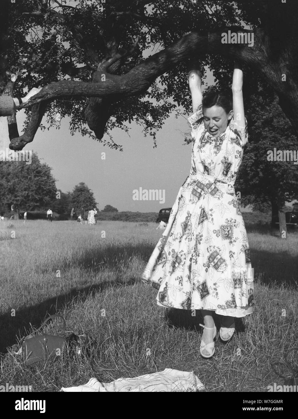 1950er Jahre, historische Yong-Frau in einem Sommerkleid, die Spaß am Ast eines Baumes im Freien im Bushy Park hat, einem großen Park rund um den Royal Palace of Hampton Court, Richmond upon Thames, London, England, Großbritannien. Ein königlicher Park, im Jahre 1529, errichtete König Heinrich VIII., ein leidenschaftlicher Jäger, die Gebiete um den Tudor-Palast als Jagdgebiet für Hirsche. Stockfoto