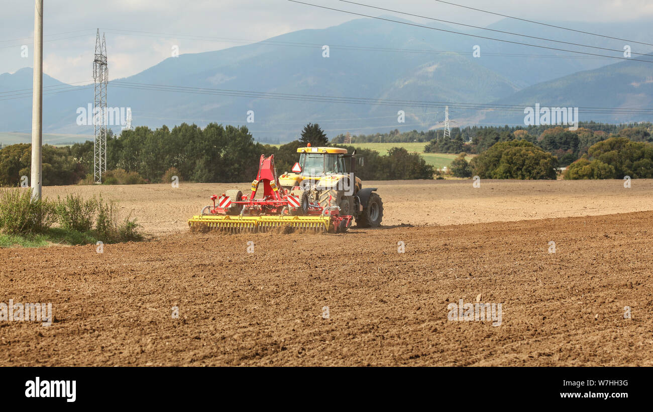 Gelbe Schlepper zieht Rot Aussaat Mechanismus über trockenes Feld, Bäume und Berge im Hintergrund Stockfoto