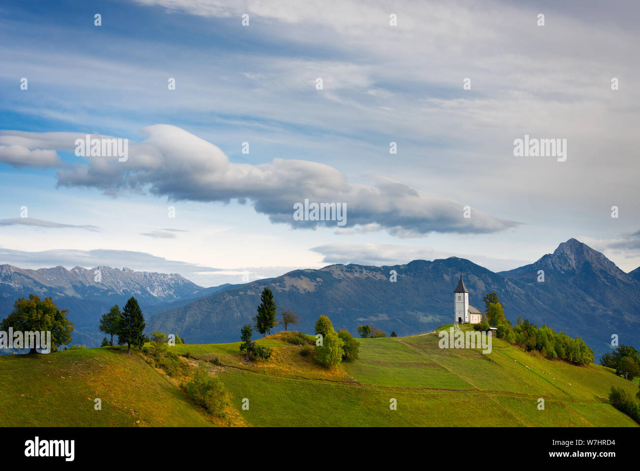 Die Kirche von St. Primus auf der Spitze eines Hügels in einer Landschaft mit der Steiner Alpen im Hintergrund unter Seltsame Wolken in Jamnik, Kranj, Slowenien. Stockfoto