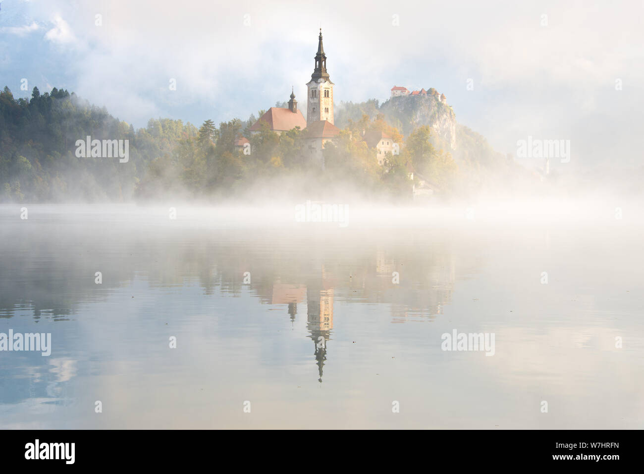Nebel über dem Wasser des Sees Bled vor der Kirche auf der Insel und im Hintergrund die Burg von Bled mit Reflexion im See in Bled, Slowenien. Stockfoto