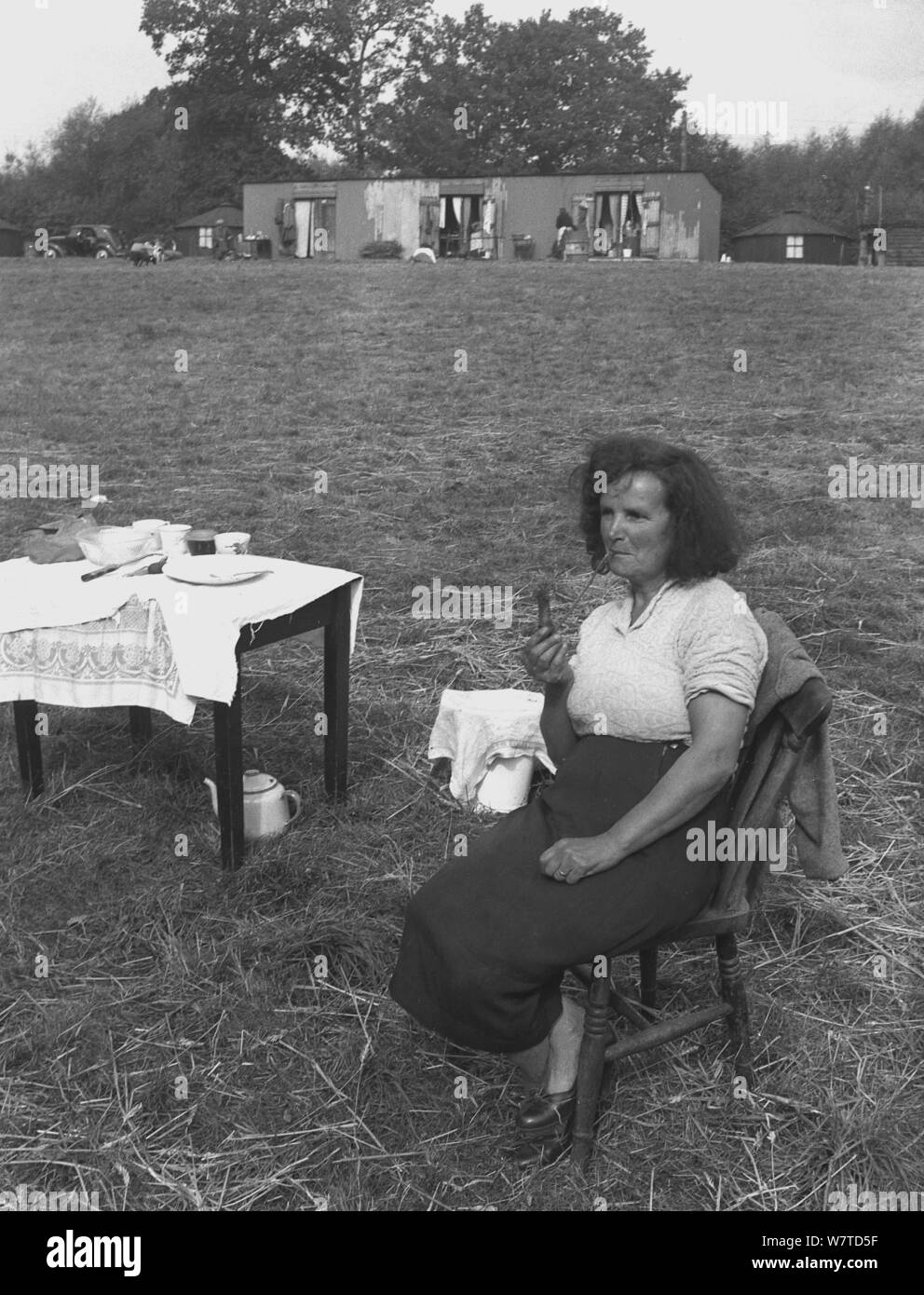 1951, historische, eine Dame landwirtschaftliche Arbeiter, ein Hop picker draußen sitzen auf einem Stuhl in einem Feld in eine Pause von der Arbeit Rauchen ein Rohr, Kent, England, UK. Das Hopper Hütten in der Ferne gesehen wurden aus Wellpappe Metall (Stahl) und waren die Unterkünfte für die Arbeiter woking in die Felder ein. Stockfoto