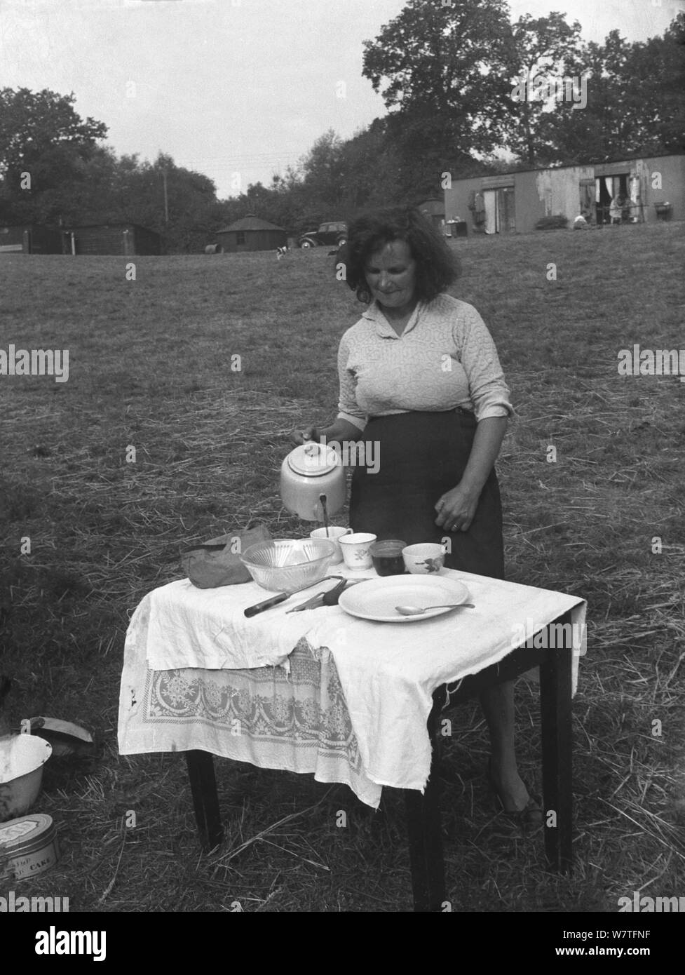 1951, historische, eine Dame landwirtschaftliche Arbeiter, ein hop Picker in einem Feld außerhalb ihrer Quartiere, genannt "Hopper Hütten", um einen kleinen Tisch mit einer Teekanne zu einer Tasse Kaffee und einem Snack, Kent, England, UK. Das Hopper Hütten in der Ferne sehen aus Wellblech gemacht wurden (Stahl) und waren die Unterkünfte für die Arbeiter woking in die Felder ein. Stockfoto