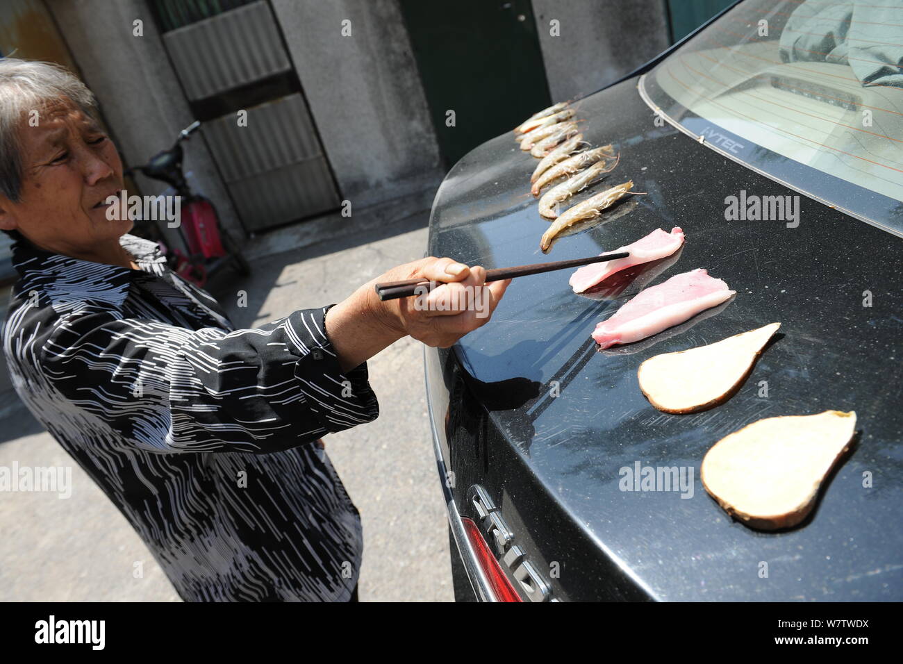 Eine ältere Frau backt Speck, Garnelen und Kartoffel auf den Kofferraum eines Autos aufgeheizt durch die sengende Sonne in Ji'nan City's East China Shandong prov Stockfoto
