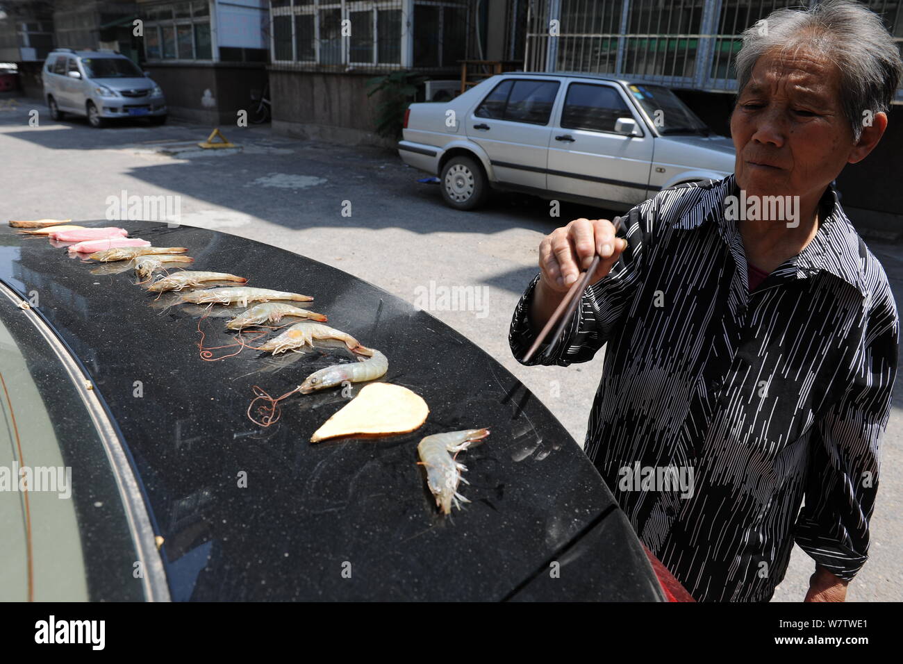 Eine ältere Frau backt Speck, Garnelen und Kartoffel auf den Kofferraum eines Autos aufgeheizt durch die sengende Sonne in Ji'nan City's East China Shandong prov Stockfoto