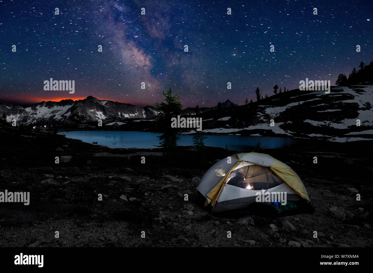 Camper innen Zelt lesen in der Nacht im Verschneiten Seen, Glühen der Waldbrand am Horizont. North Cascades Bereich des Okanogan Wenatchee National Forest, Washington, USA, Juli 2014. Model Released. Stockfoto