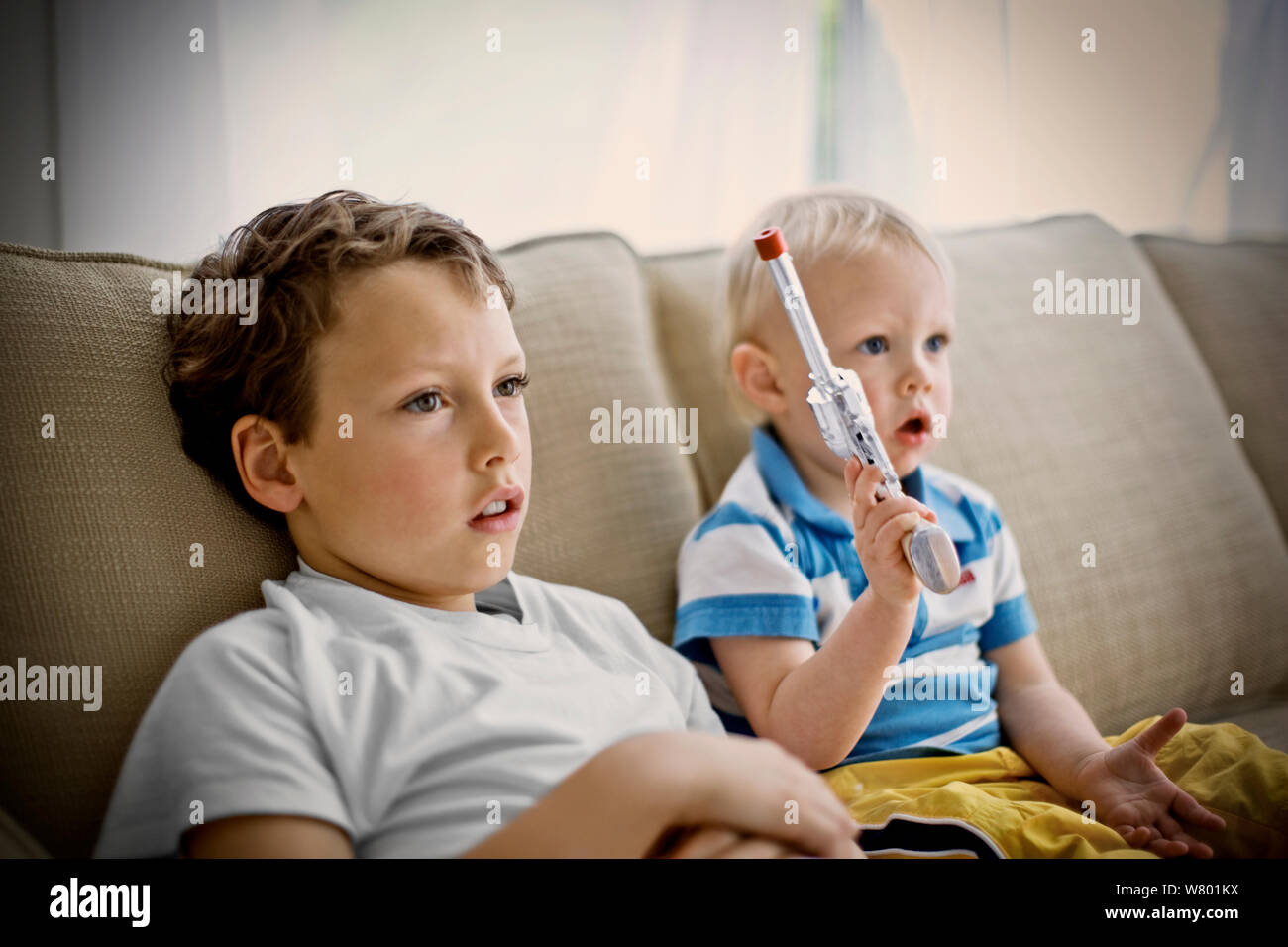 Junge sitzt auf einer Couch neben seinem jüngeren Bruder, hält eine Spielzeugpistole. Stockfoto