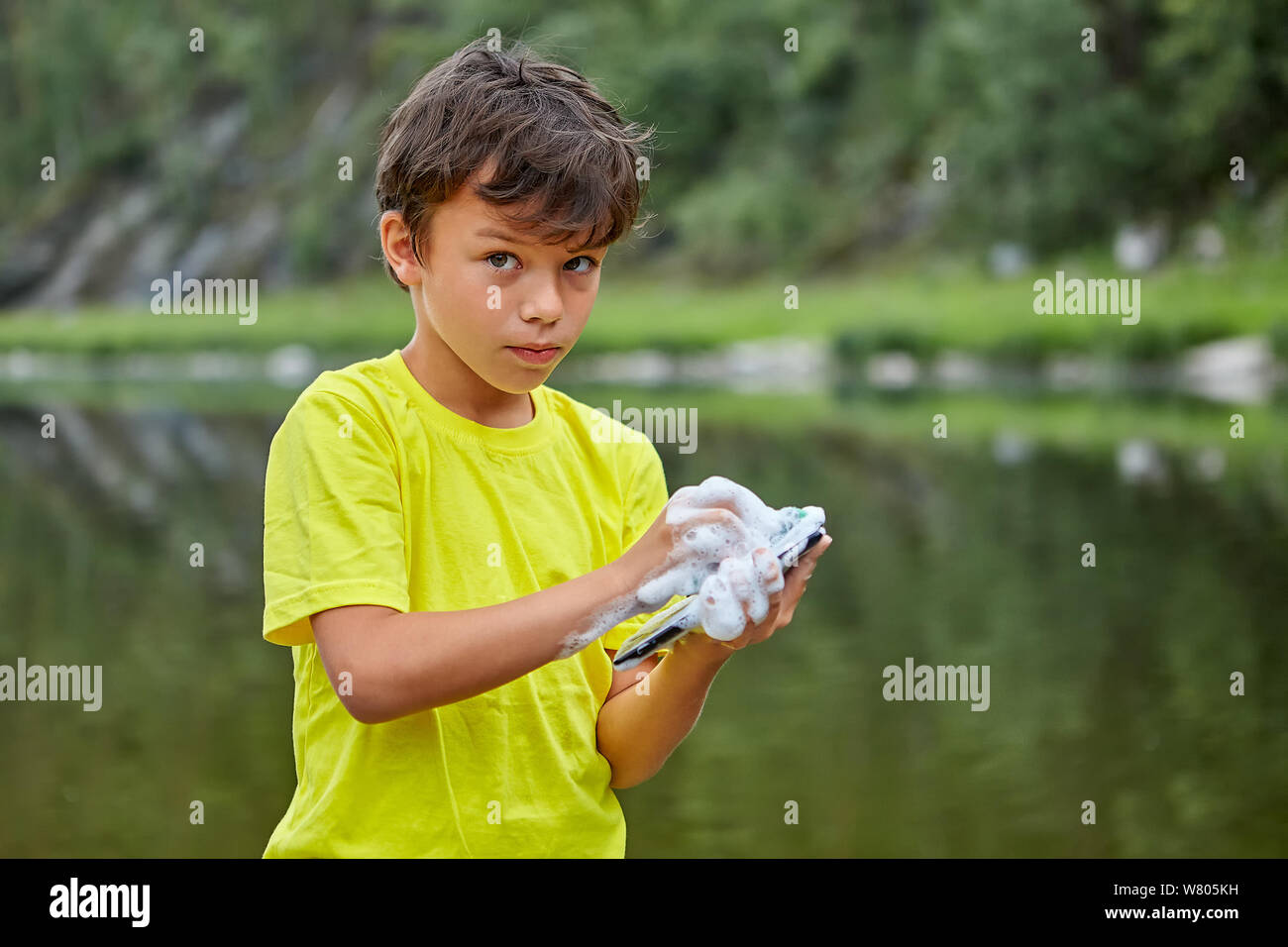 Eine ernsthafte Junge ist Waschen ein Mobiltelefon mit Seife während in der Ufer stehen. Das Kind gadget der Eltern ruiniert durch Benetzung. Stockfoto