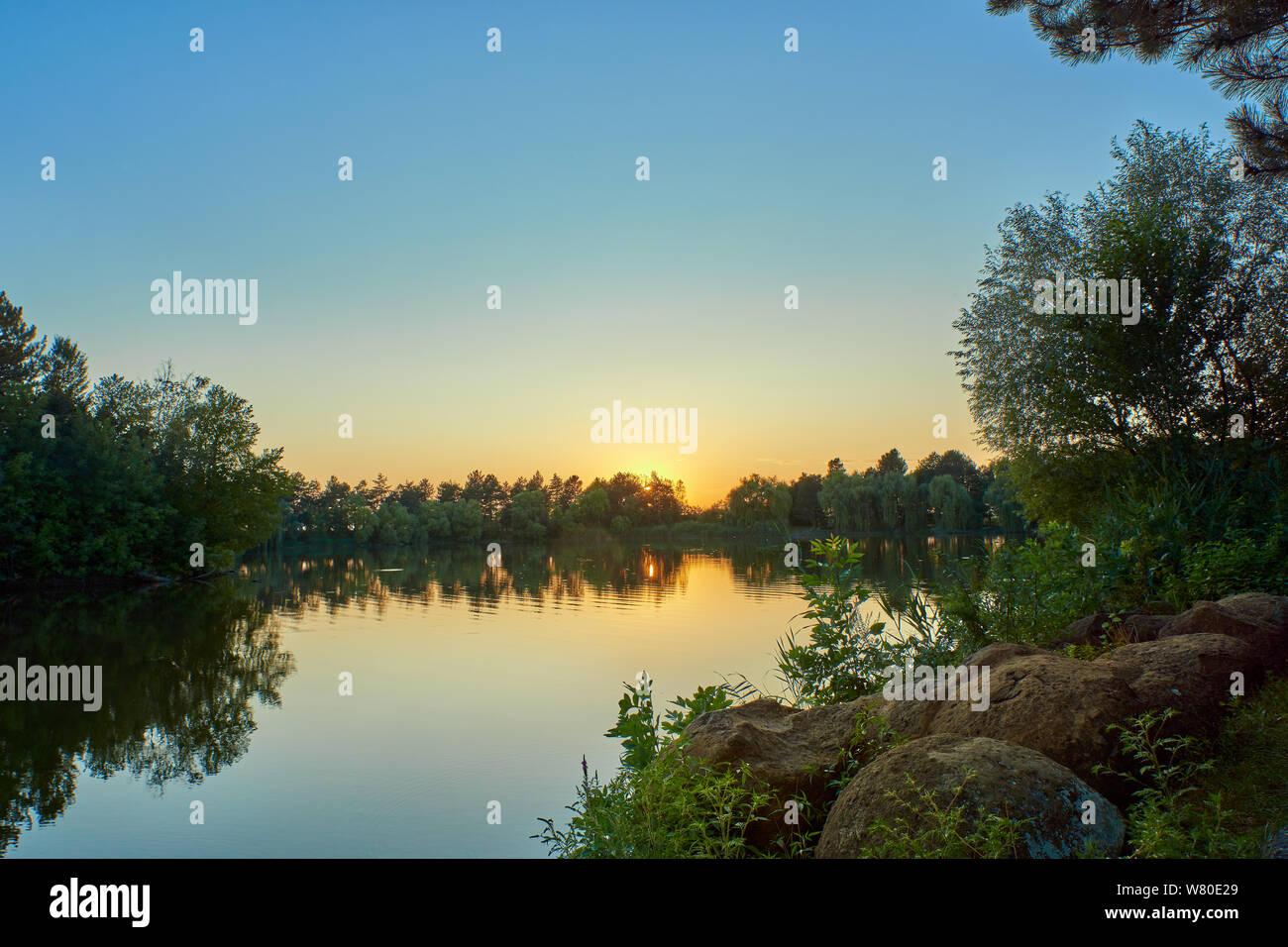 Sonnenuntergang auf einem See mit blauem Wasser und Himmel, im Vordergrund Salbei und runde Steine Stockfoto
