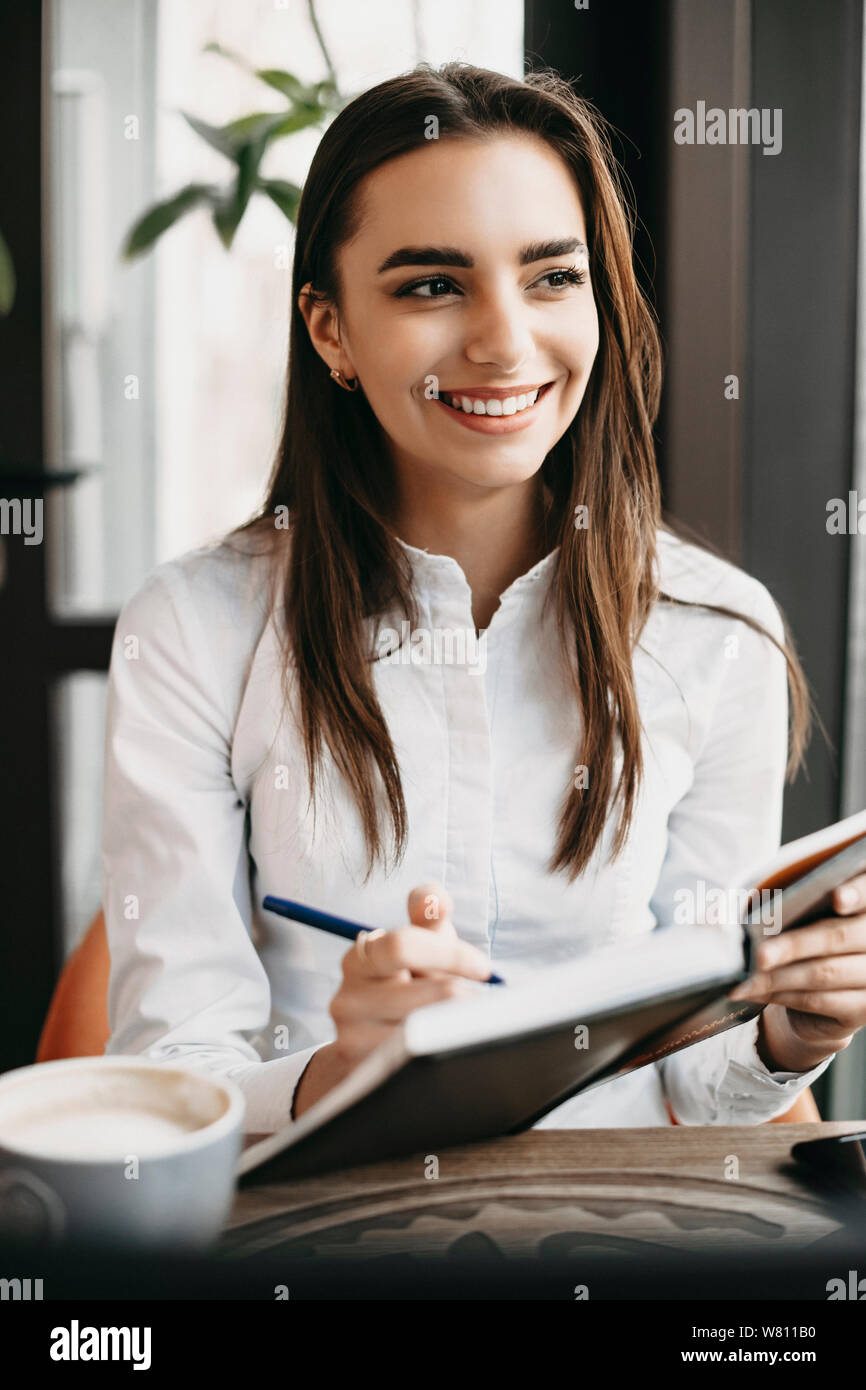 Portrait einer jungen attraktiven kaukasischen Geschäftsfrau, sitzen in einem Café am Mittag und schreiben in Ihrem Notebook weg schauen lächelnd. Stockfoto