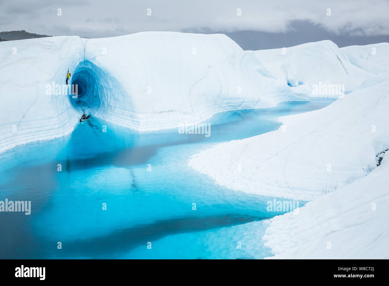 Ice climber führt eine steile Linie vor einem massiven Eishöhle in Alaska. Er hinterlässt ein Kanu mit seinem sichernde in einem blauen See Überflutung der Entra Stockfoto
