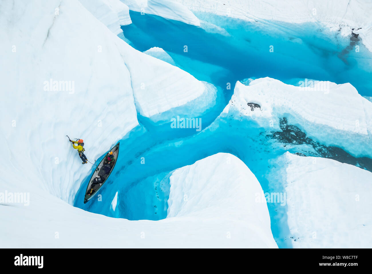 Ein ice Climber in Gelb führt ein Aufstieg nach oben und aus dem Kanu auf einem blauen See auf einem alaskischen Gletscher. Stockfoto
