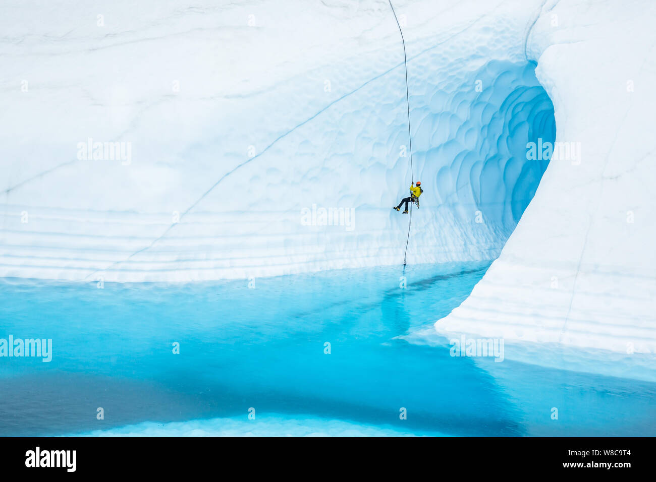 Junger Mann rappels vor einem großen Eis Höhle auf der Matanuska Gletscher. Die Höhle ist von einem lrage Blue Pool oder Supraglazialen See überflutet. Stockfoto