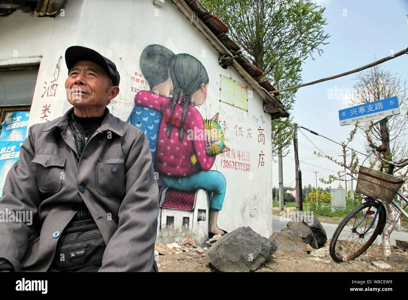 Ein Dorfbewohner sitzt vor einem Street Art Malerei des französischen Künstlers Julien Seth Malland an der Wand eines Hauses in Xinyi Dorf Fengjing Stadt in Jinsh Stockfoto