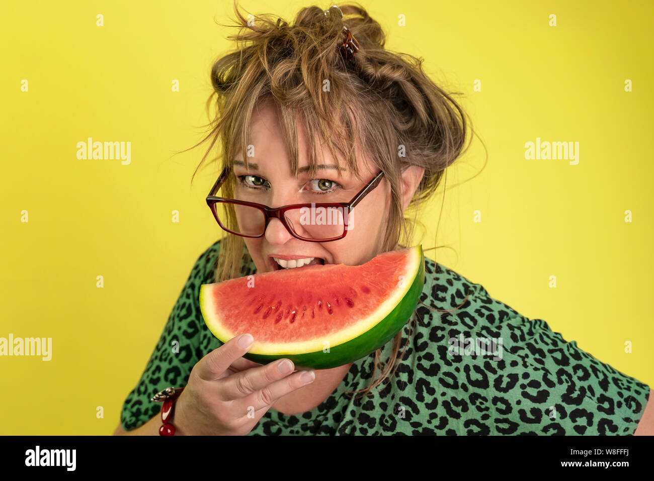 Ein Jahr 40 alte lustige Frau beißt in ein Stück Wassermelone. Sie hat wilde dunkelblonde Haare und trägt eine Brille. Gelber Hintergrund und Tier reminis Drucken Stockfoto