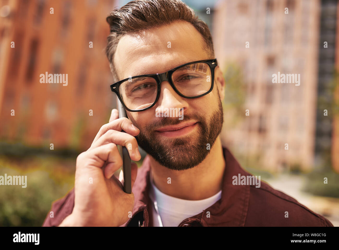 Nettes Gespräch mit Freund. Nahaufnahme Porträt von glücklichen bärtigen Mann in Casual-Kleidung und Brillen sprechen per Telefon und lächeln, während auf der Straße stehen. Lifestyle. Digital. Kommunikation Stockfoto