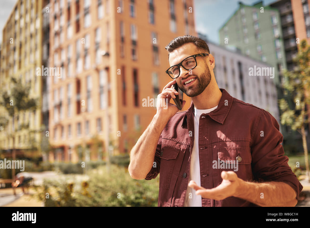 Gute Nachrichten glücklicher bärtiger Mann in Casual Wear und Brillen telefonieren und lächeln, während er auf der Straße steht. Lifestyle. Digital. Kommunikation Stockfoto