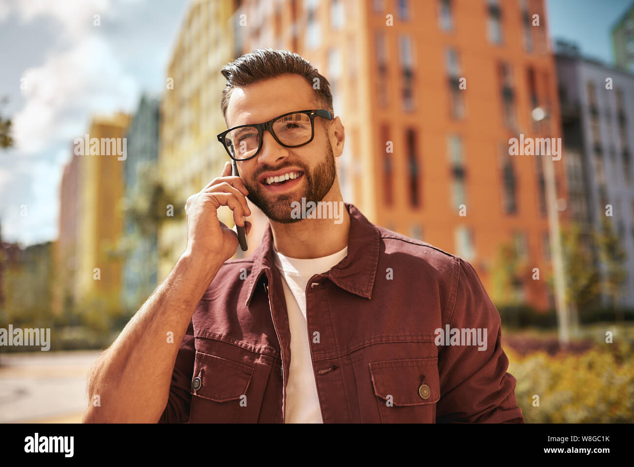 Glücklich, Sie zu hören Porträt von fröhlichen und gut aussehenden bärtigen Mann in Freizeitkleidung und Brillen sprechen per Telefon und lächeln, während auf der Straße stehen. Positive Emotionen. Digital. Kommunikation Stockfoto