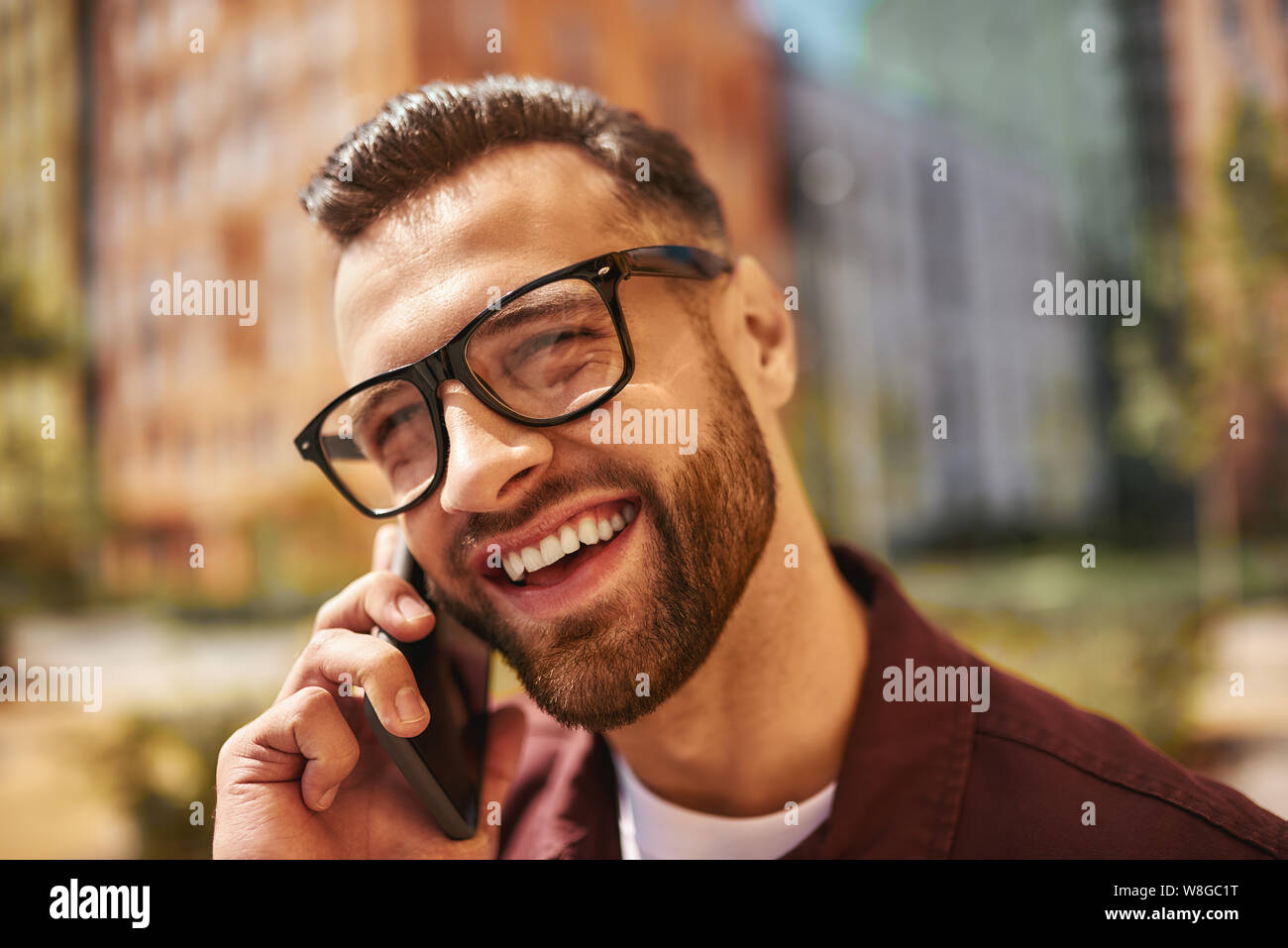 Wir teilen großartige Neuigkeiten. Nahaufnahme Porträt von glücklichen bärtigen Mann in Casual-Kleidung und Brillen sprechen per Telefon und lächeln, während auf der Straße stehen. Positive Emotionen. Digital. Kommunikation Stockfoto