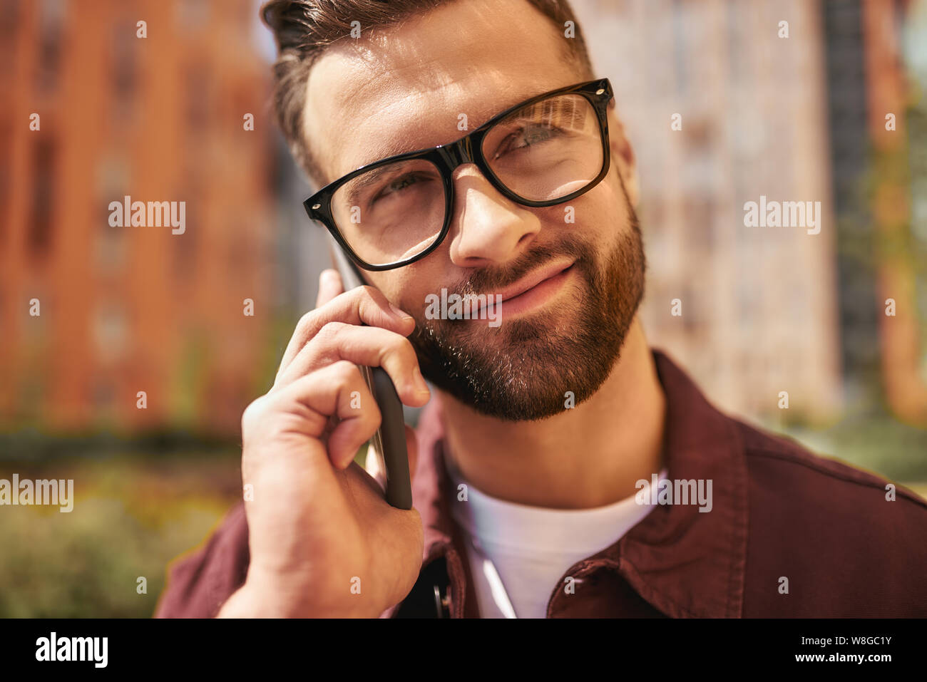 Wir teilen großartige Neuigkeiten. Nahaufnahme Porträt von glücklichen bärtigen Mann in Casual-Kleidung und Brillen sprechen per Telefon und lächeln, während auf der Straße stehen. Lifestyle. Digital. Kommunikation Stockfoto