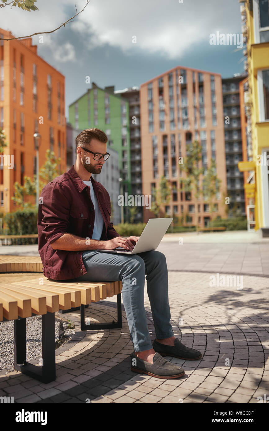 Guter Arbeitstag. In voller Länge von jungen schönen Mann mit Stoppeln in Freizeitkleidung und Brillen arbeiten auf Laptop, während auf der Bank im Freien sitzen. Bloggen. Arbeitskonzept. Stadtleben Stockfoto