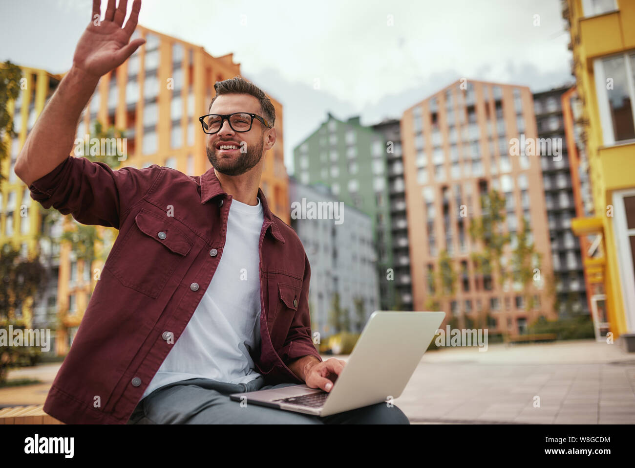 Ich bin hier Junge und fröhlich schöner Mann mit Stoppeln in Freizeitkleidung und Brille winkt einem Freund, während er auf der Bank mit Laptop im Freien sitzt. Begrüßung. Bloggen. Stadtleben Stockfoto