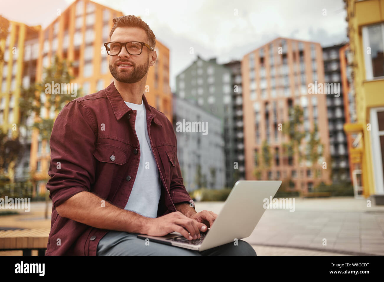 Arbeiten im Freien. Junger gutaussehender Mann mit Stoppeln in legerer Kleidung und Brillen, die am Laptop arbeiten, während sie draußen auf der Bank sitzen. Bloggen. Stadtleben Stockfoto