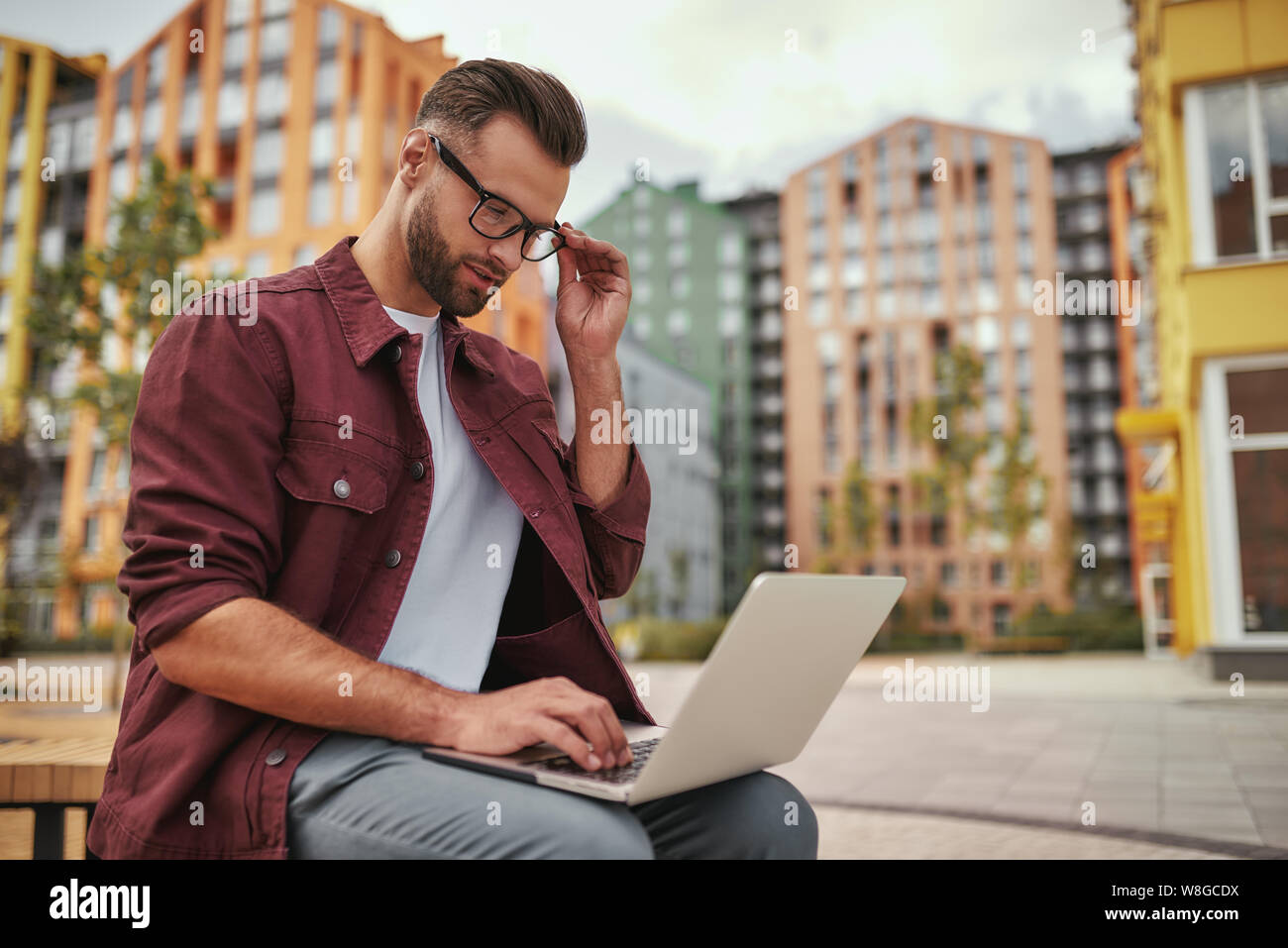 E-Mail wird geprüft Junger, hübscher Mann mit Stoppeln in legerer Kleidung, der Brillen justiert und am Laptop arbeitet, während er auf der Bank im Freien sitzt. Bloggen. Arbeitskonzept. Stadtleben Stockfoto