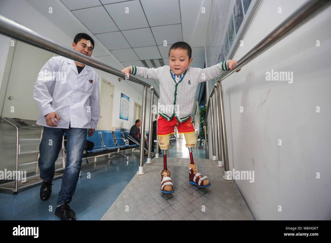 Die 4-jährige Beinlose junge Xiao Feng Praktiken gehen mit Hilfe von temporären prothetischen Beine neben einem Physiotherapeuten an der Behinderte Rehabili Stockfoto