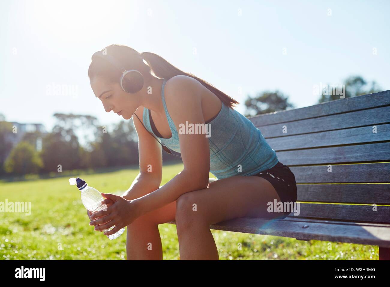 Junge Frau mit Kopfhörern sitzt auf der Bank mit einer Flasche Wasser. Stockfoto
