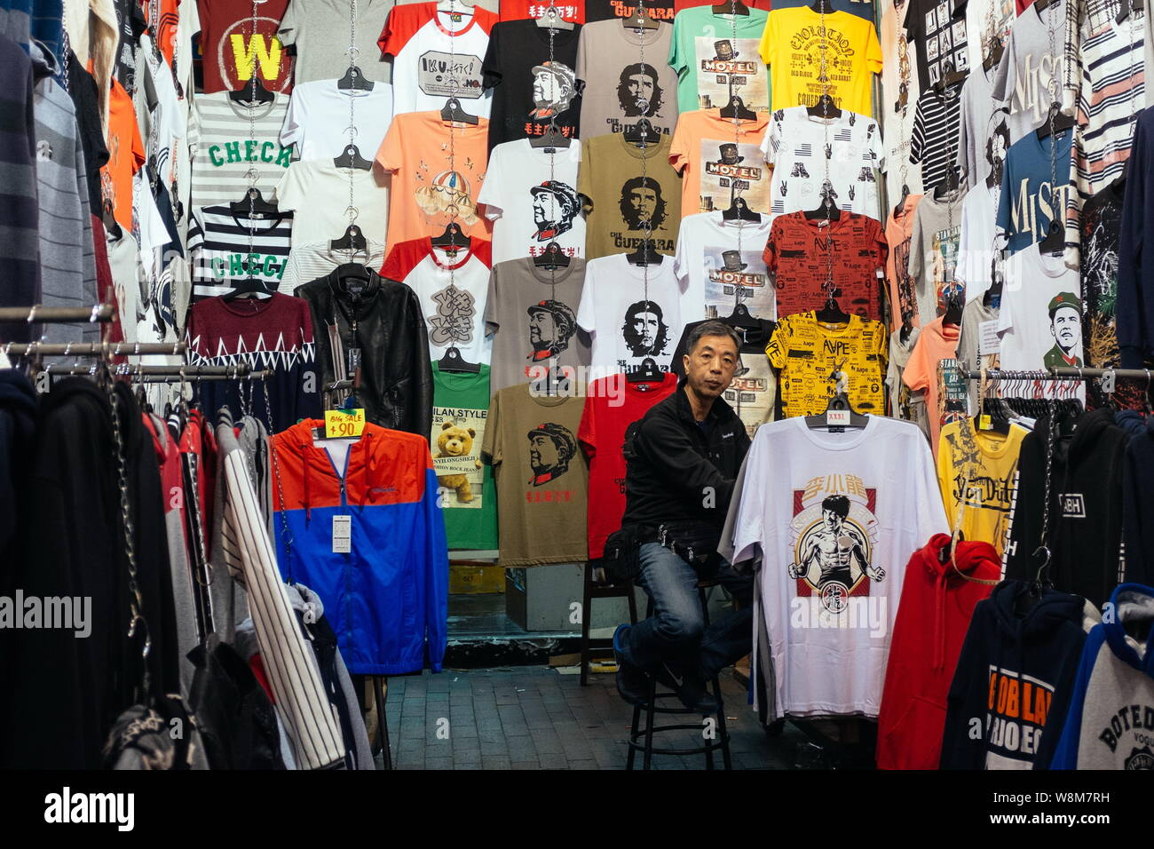 Ein Anbieter Verkauf von Che Guevara, Bruce Lee, Mao Zedong und andere T-Shirts sitzt an seinem Stall in einem behelfsmäßigen Nachtmarkt in der Temple Street in Hongkong, Ch Stockfoto