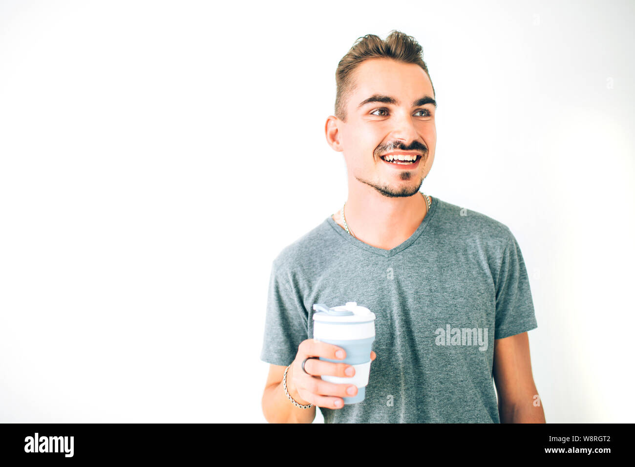 Glückliche junge Mann im grauen T-Shirt mit einer Tasse Kaffee auf weißem Hintergrund isoliert. Weg schauen lächelnd. Stockfoto