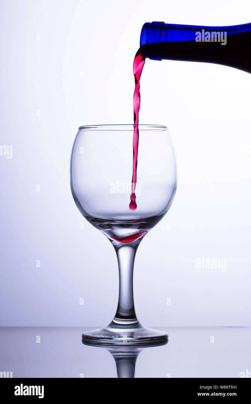 Der erste Tropfen fällt in ein leeres Glas. Ein Jet der roten Wein fließt in ein Glas aus einer blauen Flasche. Wein Jet nicht das Glas erreichen. Stockfoto