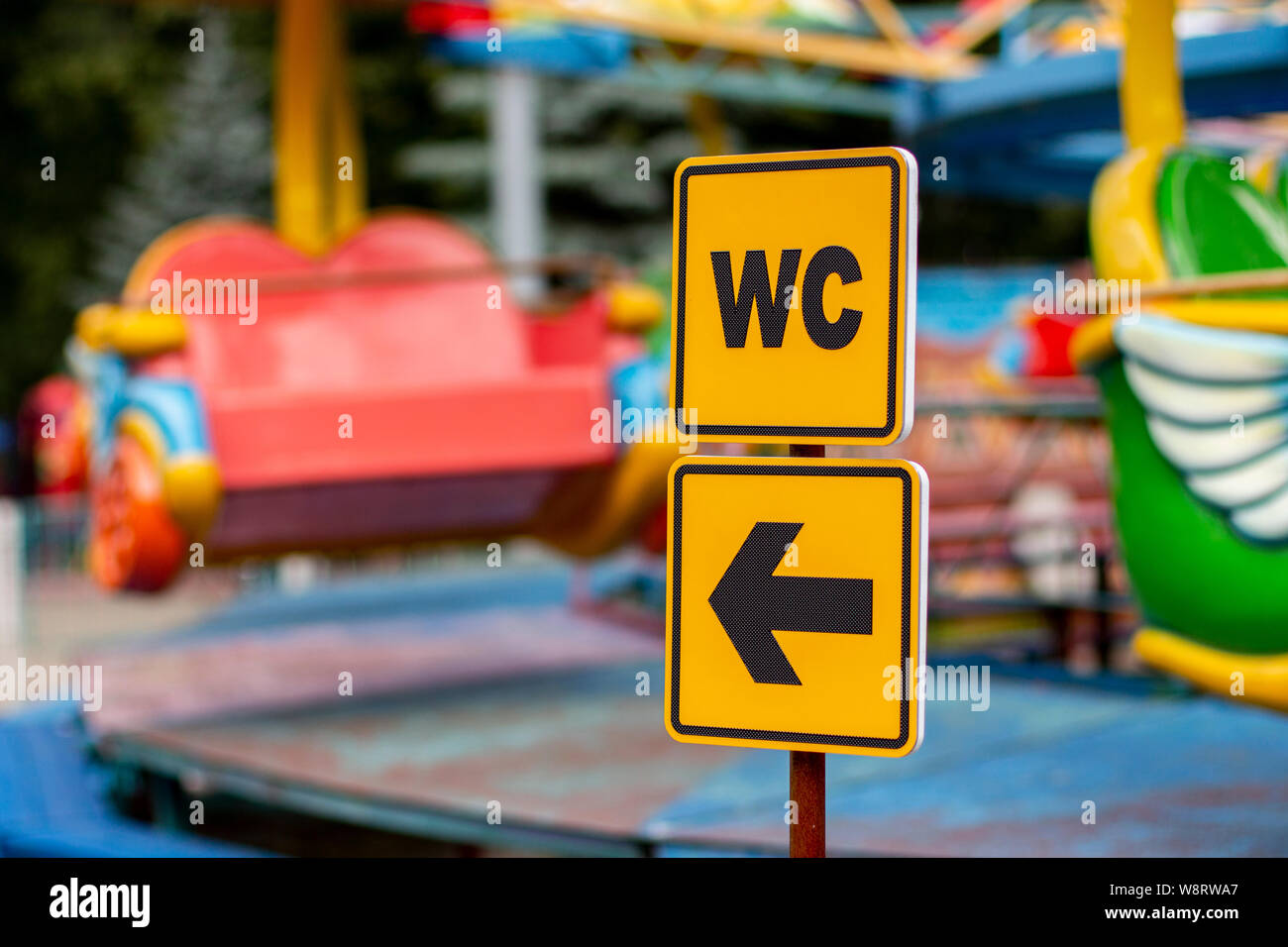 WC-Schild auf ein gelbes Schild mit Pfeil in einem öffentlichen Vergnügungspark. Zeichen Zeiger, wo die Toilette befindet, horizontale Unschärfe-Effekte im Hintergrund Stockfoto