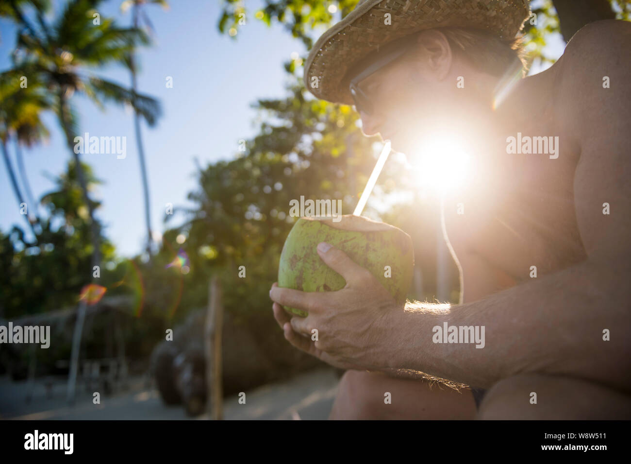 Mann mit einem Stroh Sonnenhut mit einem grünen Kokosnuss Hintergrundbeleuchtung von tropischen Sonne auf einer abgelegenen Insel Strand Stockfoto