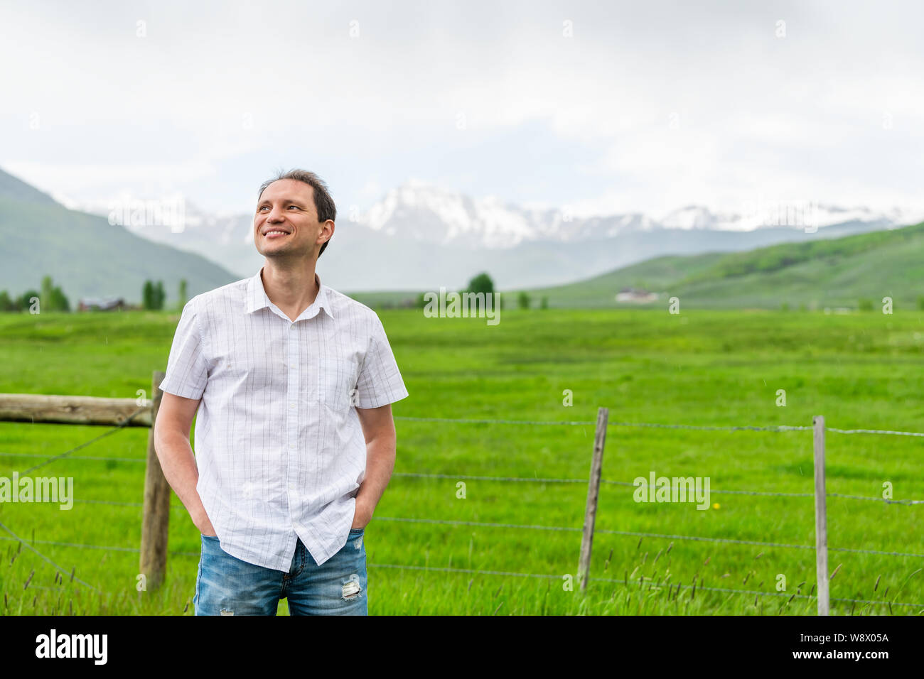 Crested Butte, Colorado Land mit glücklichen jungen Mann durch Zaun und im Sommer auf bewölkten Tag mit grünem Gras und Blick auf die Berge Stockfoto