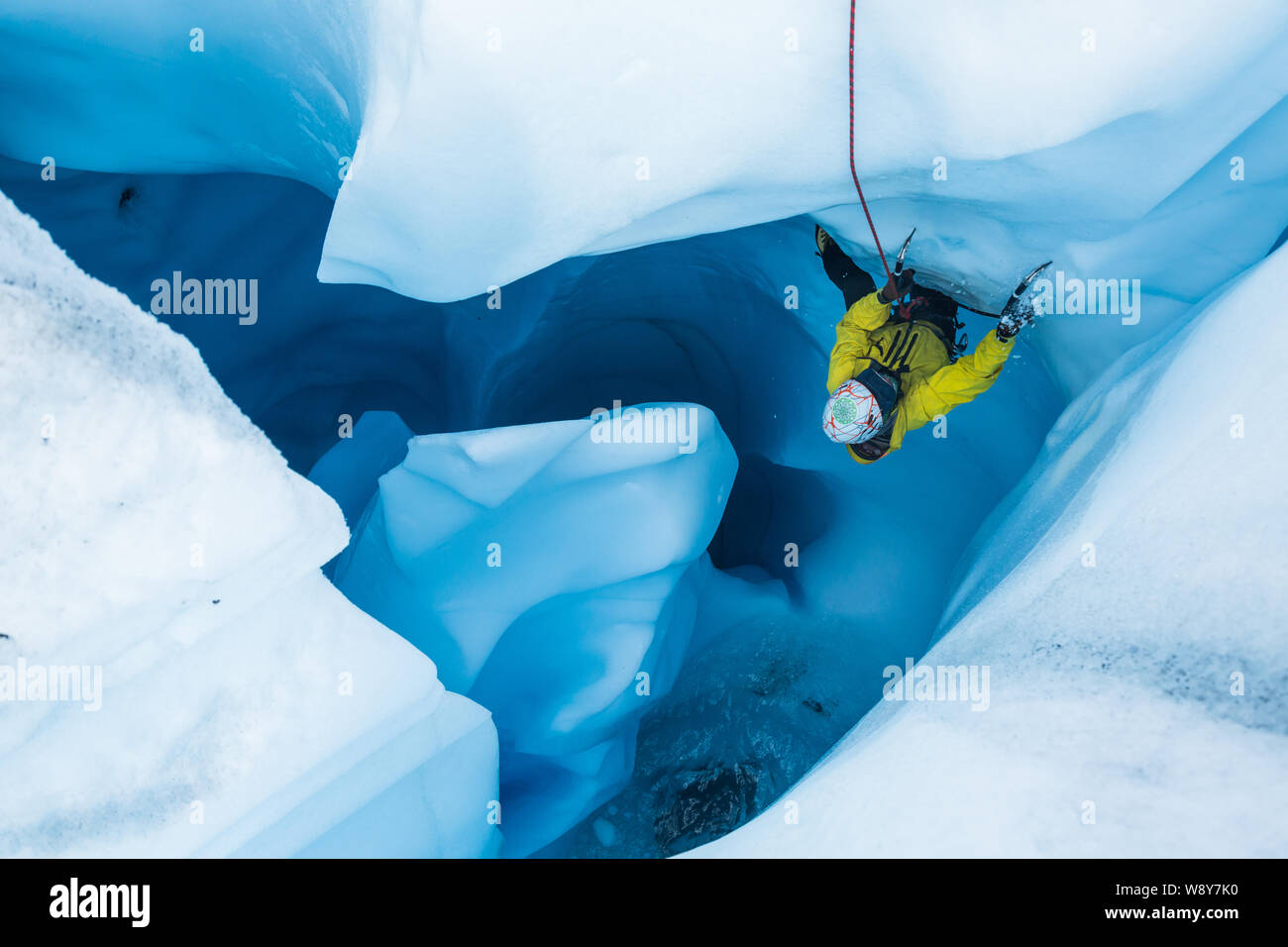 Mann in Gelb jecket Klettern eine überhängende Teil des Eises in einem Gletscher Moulin in Alaska. Seinen Fuß aus NACHLASSENDER als sein Werkzeug bricht das Eis in kleine Stockfoto