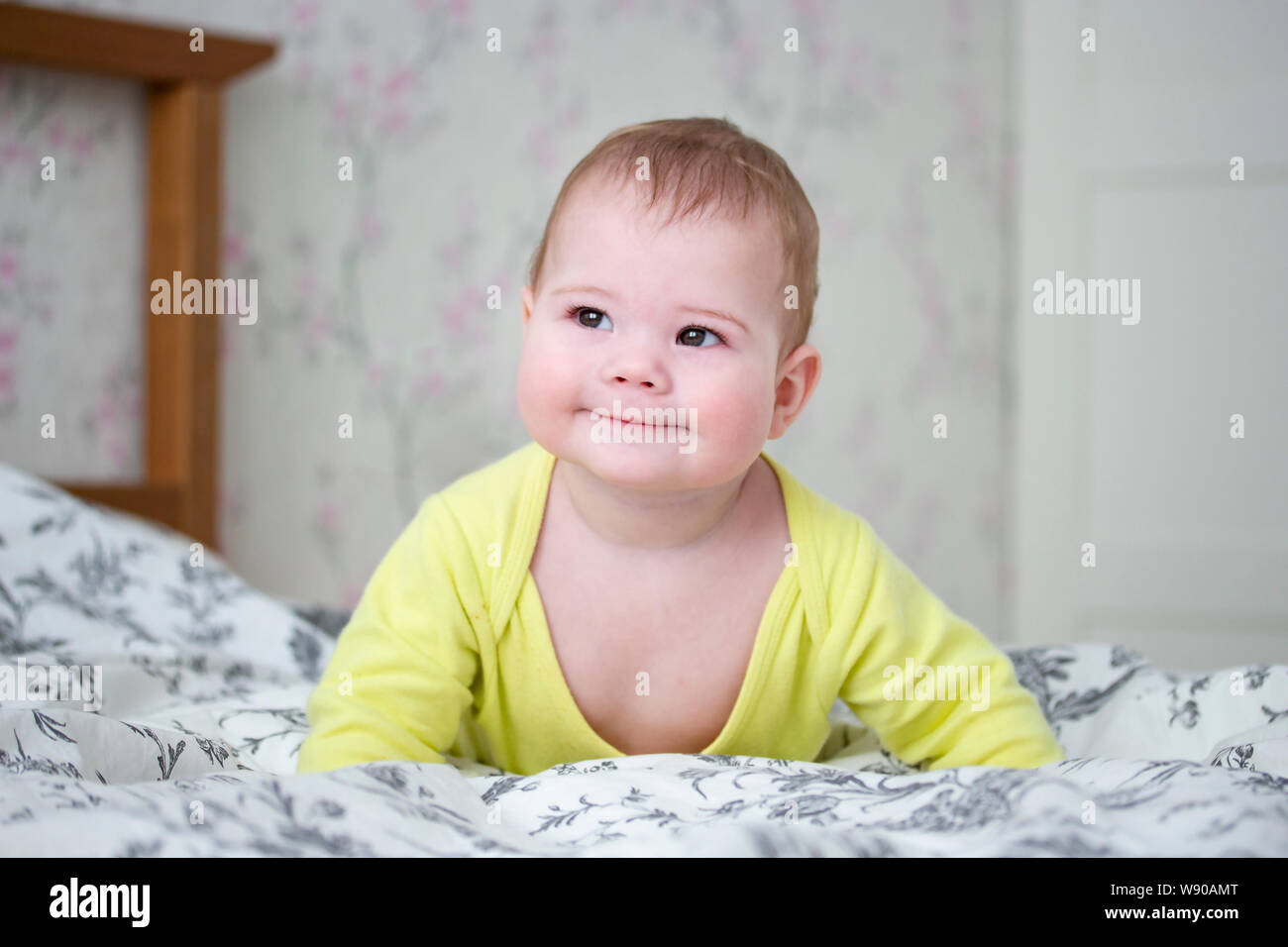 Wenig Europäische 7 Monate Baby Mädchen Jungen in Gelb. Niedliche Kind wirft seinen Körper in seinen Armen, lächelt verschmitzt, Schiele seine Augen, liegt auf dem Bett Stockfoto
