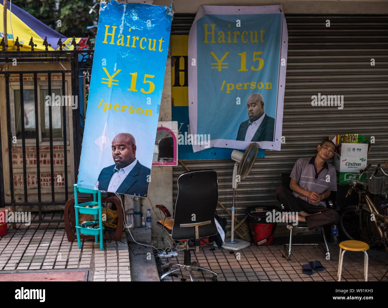 Ein chinesischer Friseur sitzt neben Plakate zeigen Haarschnitt Preise und das Bild von einem afrikanischen Mann an seiner in der Schokolade Stadt in Guangzhou City ausgeht, Sou Stockfoto