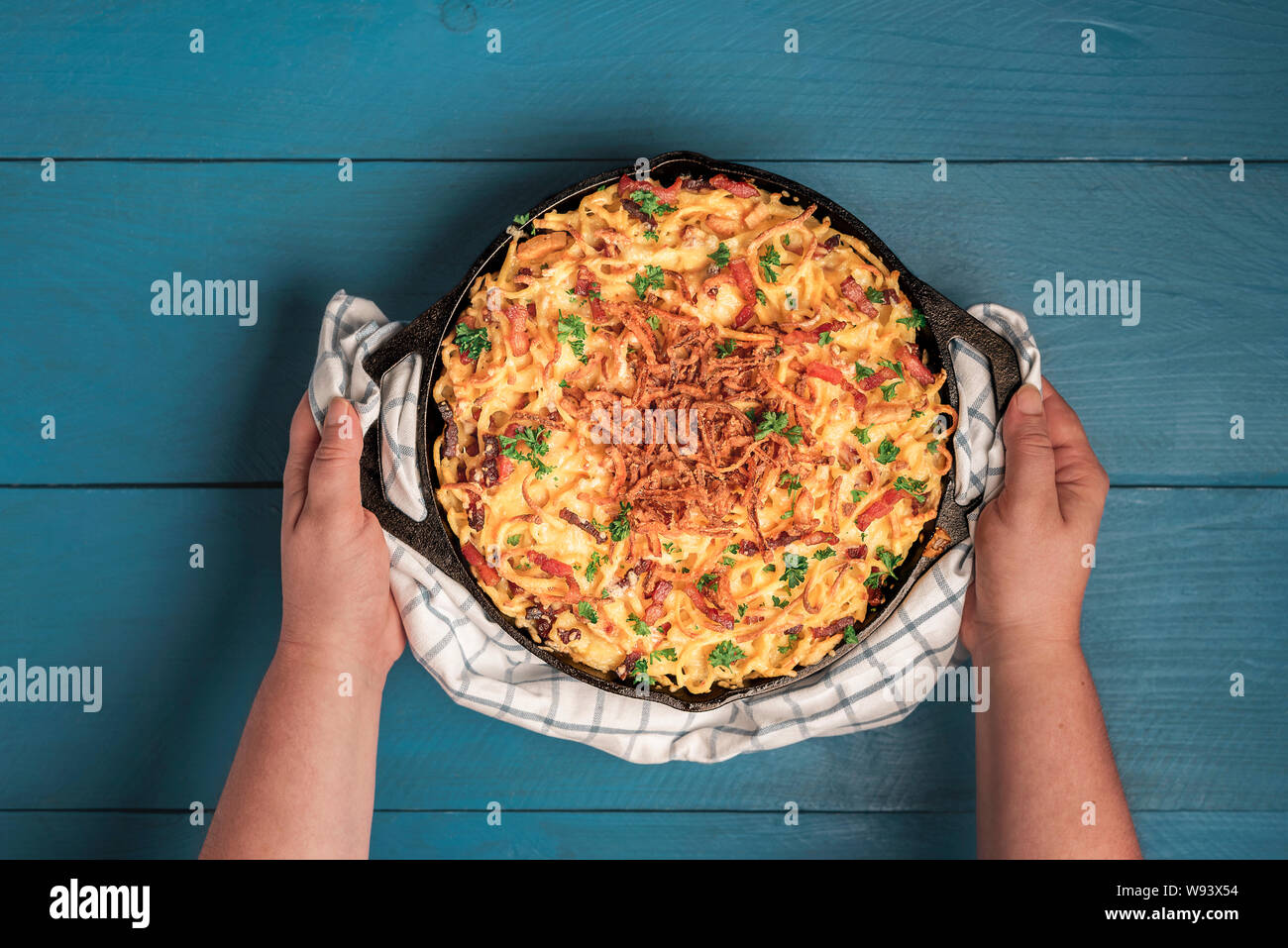 Frau Hände halten frisch zubereitete deutsche Käse Pasta über blau Holztisch. Flach der Schwäbischen traditionelle Nudeln mit Käse und Speck. Stockfoto