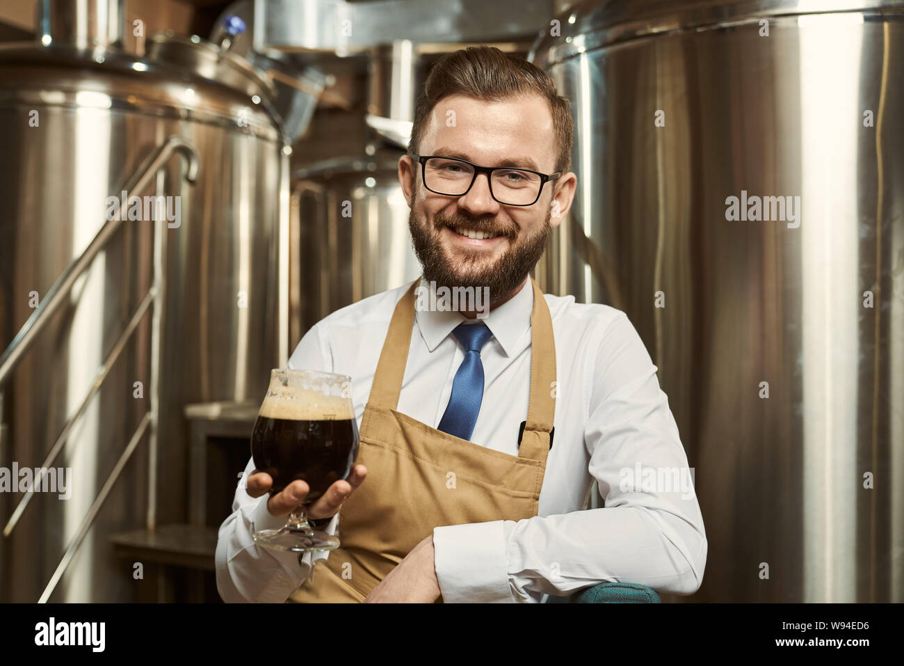 Vorderansicht der freundliche Mann mit weißem Hemd und Schürze auf Kamera und lachend, während Sie ein Glas Bier. Professionelle Brauer genießen Leckere dunkle Ale. Begriff der Produktion. Stockfoto